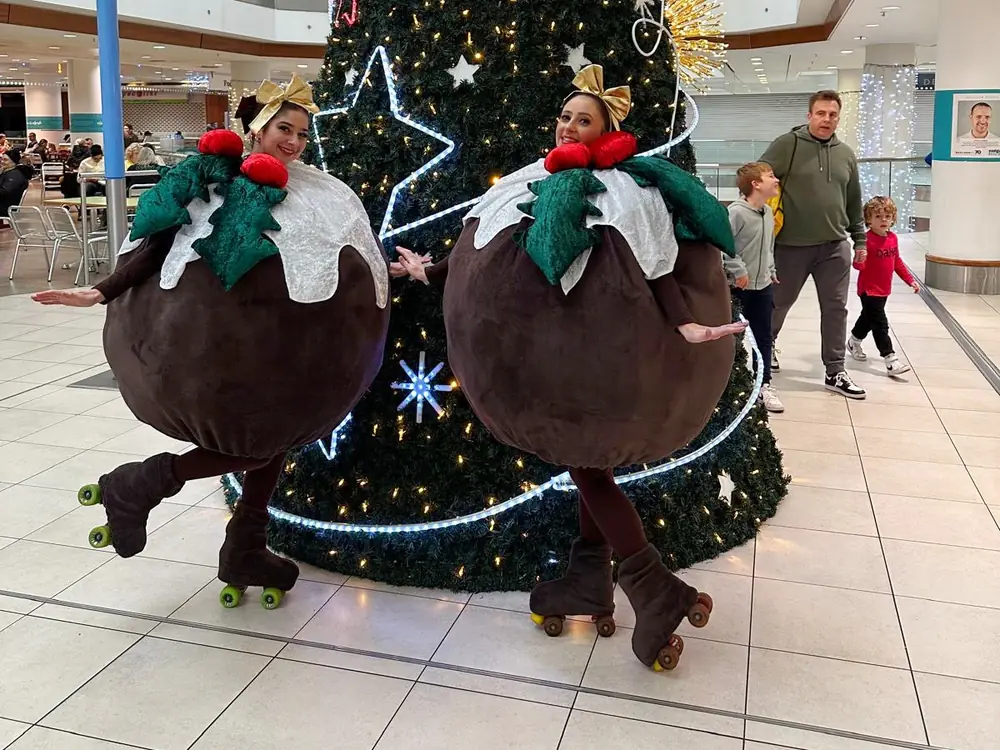 Two roller skating Christmas pudding performers beside a giant Christmas tree in a shopping centre