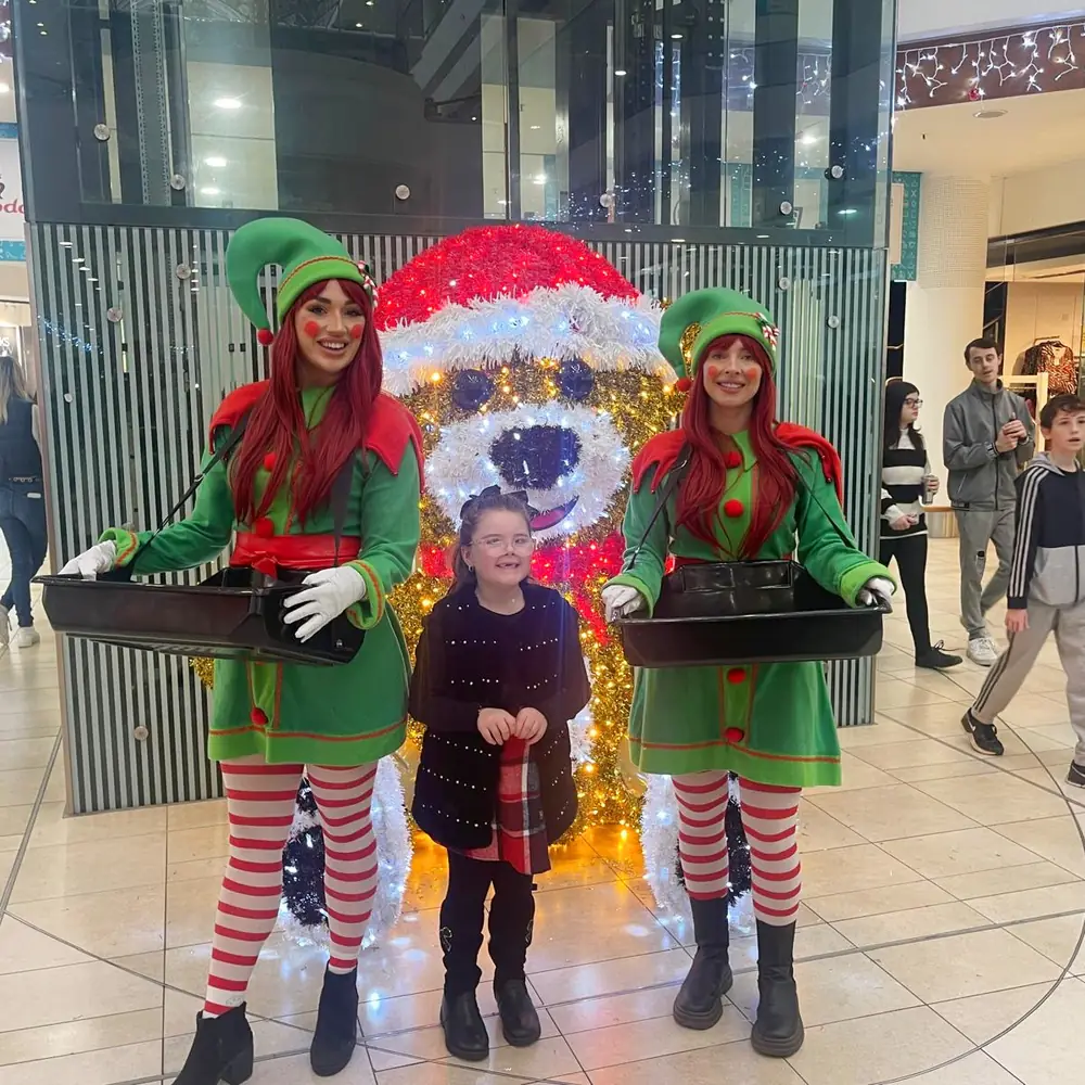Two Christmas elf performers posing with a young girl in front of a light-up bear in a shopping centre