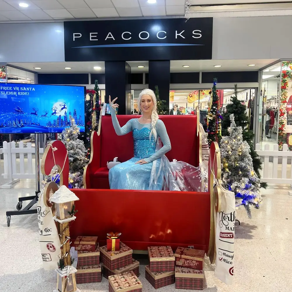 Ice princess character waving from a red Santa sleigh outside Peacocks in a shopping centre